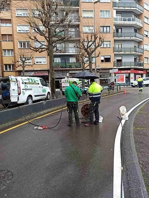 Los trabajadores limpian la calle mojada junto a los edificios de apartamentos después de un incidente, con una furgoneta y barreras cerca.