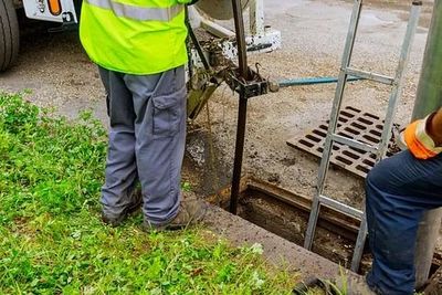 Trabajadores en una zanja al borde de la carretera; uno de ellos lleva un chaleco reflectante junto a una escalera metálica y una rejilla de drenaje.