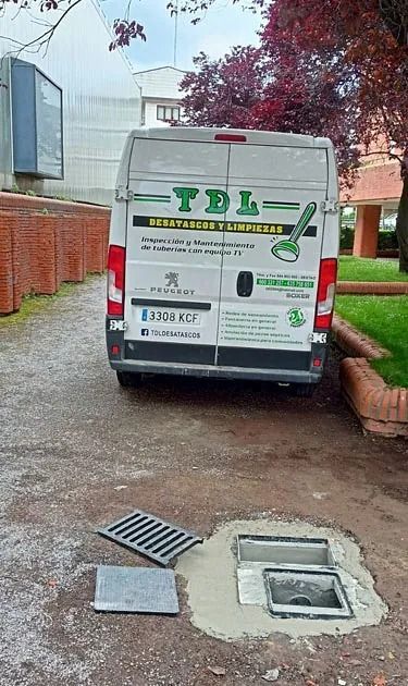 Furgoneta blanca de servicio estacionada en un camino de tierra junto a un muro de ladrillos, con una rejilla de drenaje y un acceso abierto para servicios públicos en primer plano.