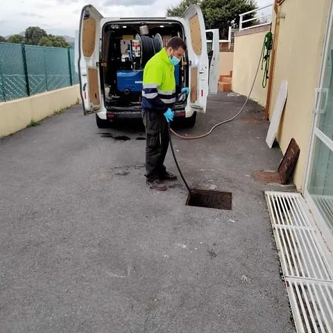 Un trabajador con chaleco de seguridad amarillo desatasca un desagüe con una manguera junto a una furgoneta de servicio.