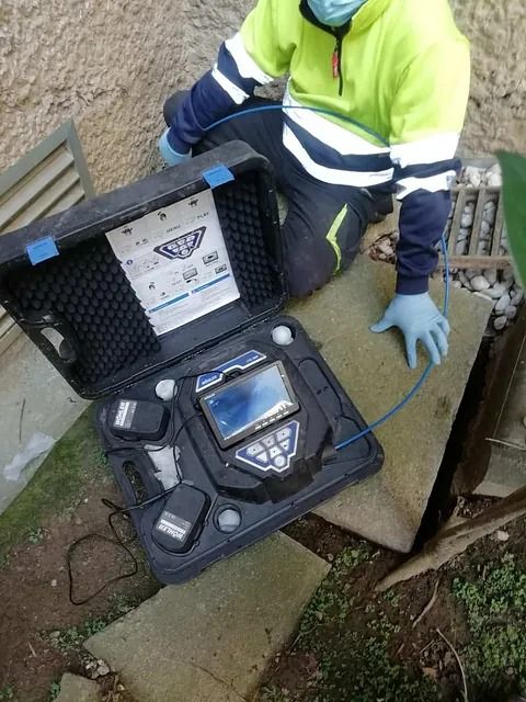 Un trabajador con chaleco reflectante se arrodilla junto a una caja de equipos abierta con un dispositivo y herramientas sobre escalones de piedra al aire libre.