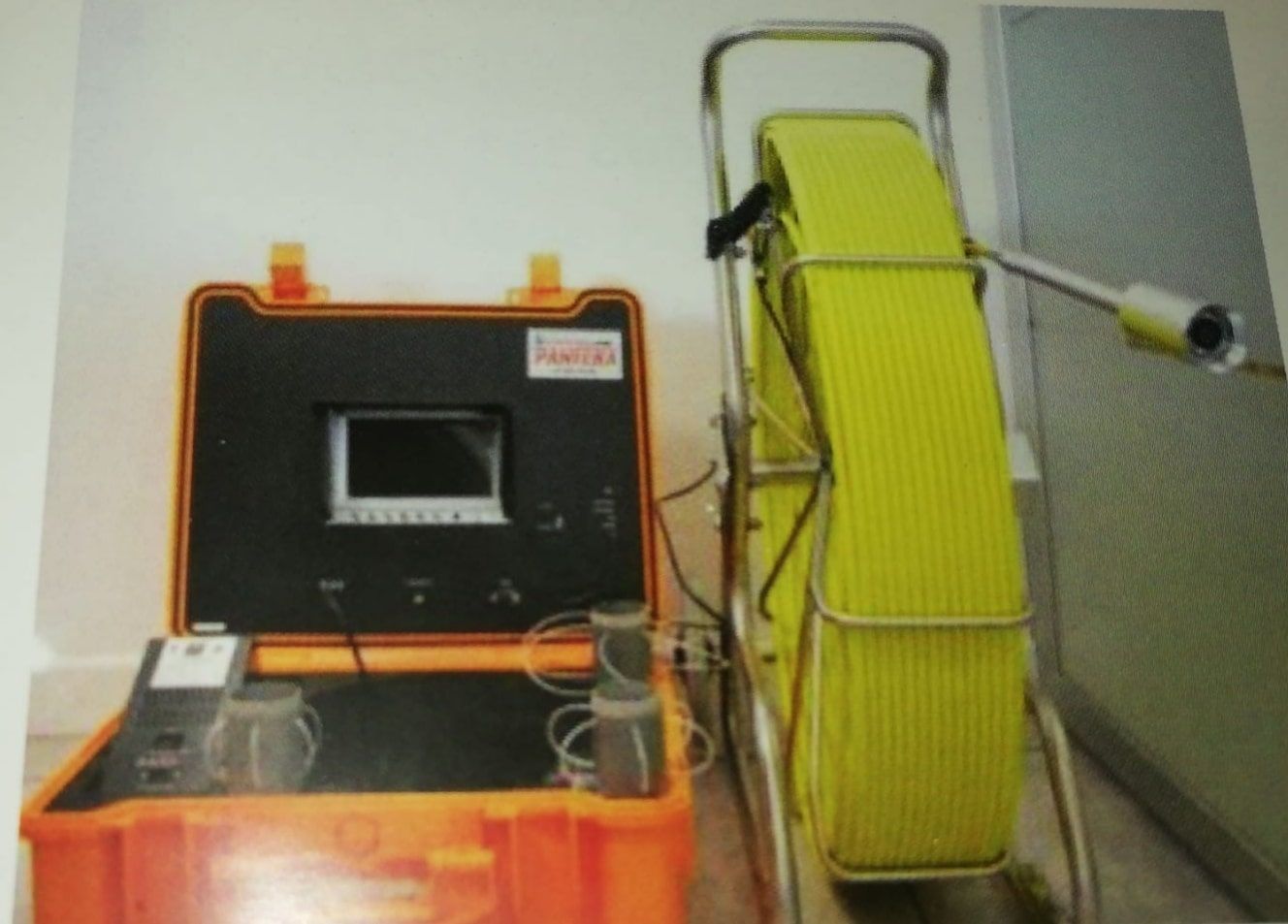 Máquina negra con pantalla pequeña junto a un carrete de manguera amarillo en un laboratorio/área de trabajo interior.