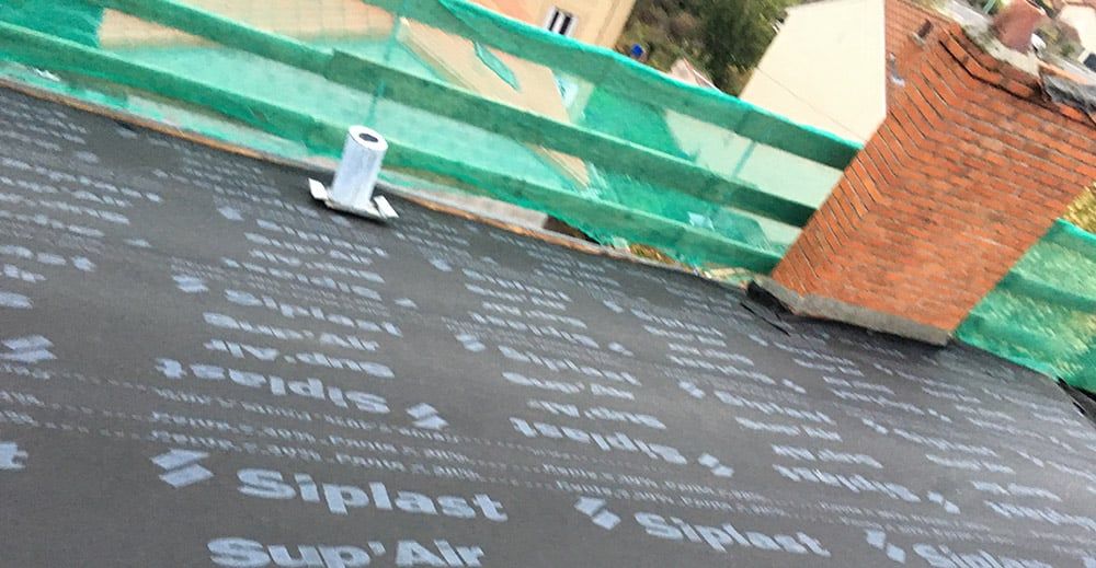 Toiture noire avec logo « Siplast » sur un toit avec cheminée et tuyau. Filet de sécurité vert.