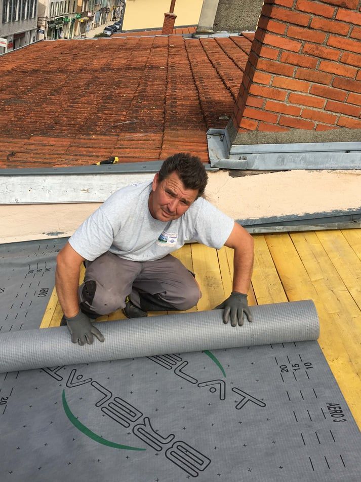 Un homme agenouillé sur un toit déroule un matériau de toiture gris ; cheminée en briques en arrière-plan.
