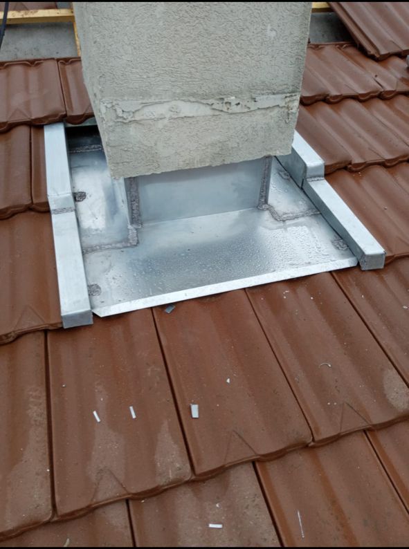 Gros plan d'une base de cheminée avec solin métallique sur un toit en tuiles brunes.