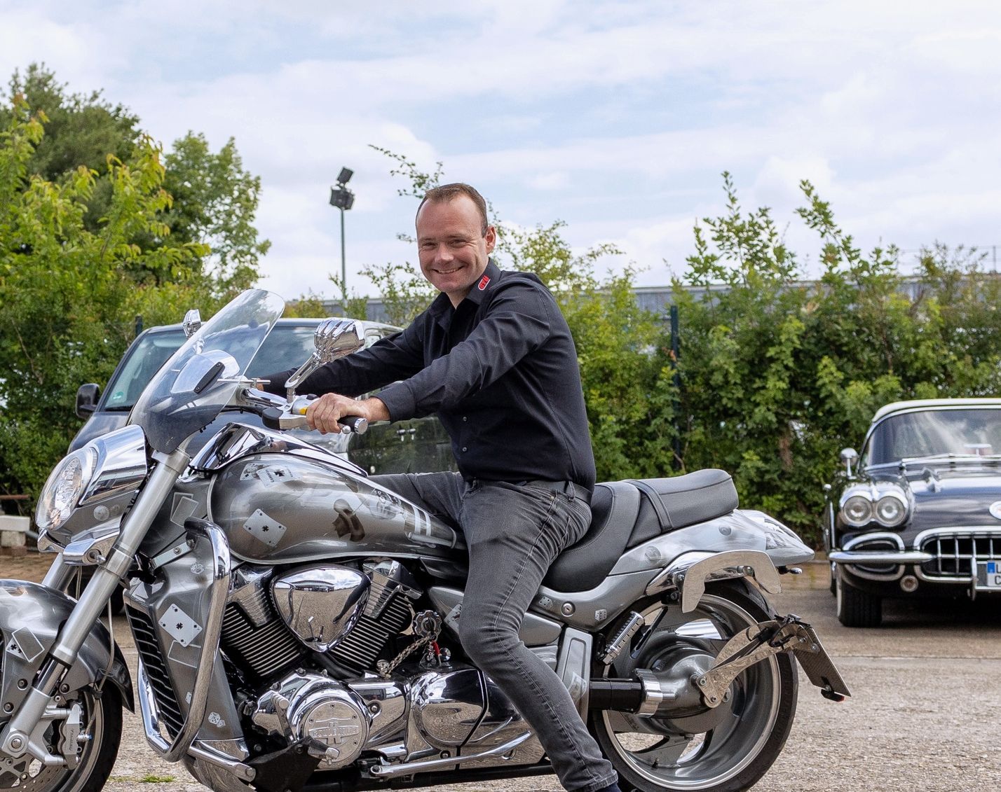 Mann sitzt lächelnd auf einem silbernen Motorrad. Draußen auf einem Parkplatz.