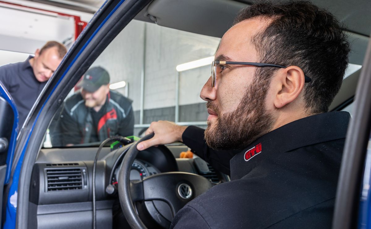 Mann fährt Auto in einer Werkstatt, zwei Mechaniker schauen sich das Fahrzeug an.