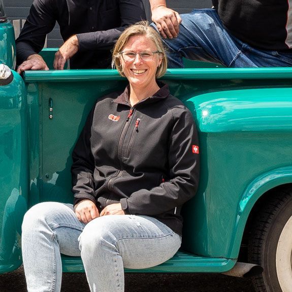 Eine Frau in einer schwarzen Jacke und Jeans sitzt lächelnd auf der Ladefläche eines blaugrünen Pickups.