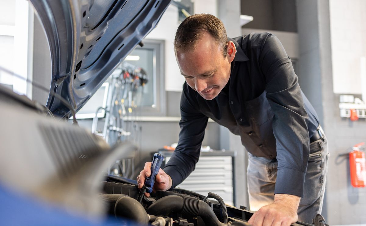 Mechaniker überprüft in einer Werkstatt einen Automotor mit einer Taschenlampe; konzentrierter Gesichtsausdruck.
