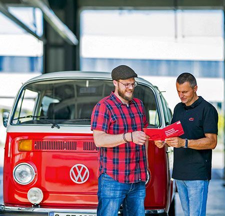 Zwei Männer untersuchen eine rote Broschüre neben einem alten roten Volkswagen-Bus in einer Garage.