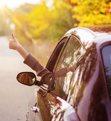 Person in einem kastanienbraunen Auto zeigt den Daumen nach oben, Arm aus dem Fenster; helles Sonnenlicht.