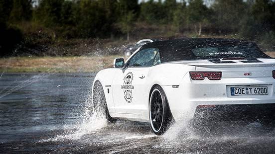 Weißes Cabrio, das auf einer Rennstrecke durchs Wasser spritzt, schwarze Räder, dunkles Verdeck.