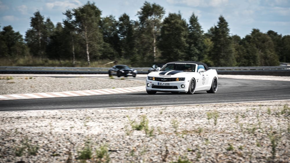 Weißes Cabrio auf der Rennstrecke, das ein schwarzes Auto verfolgt. Bäume im Hintergrund.