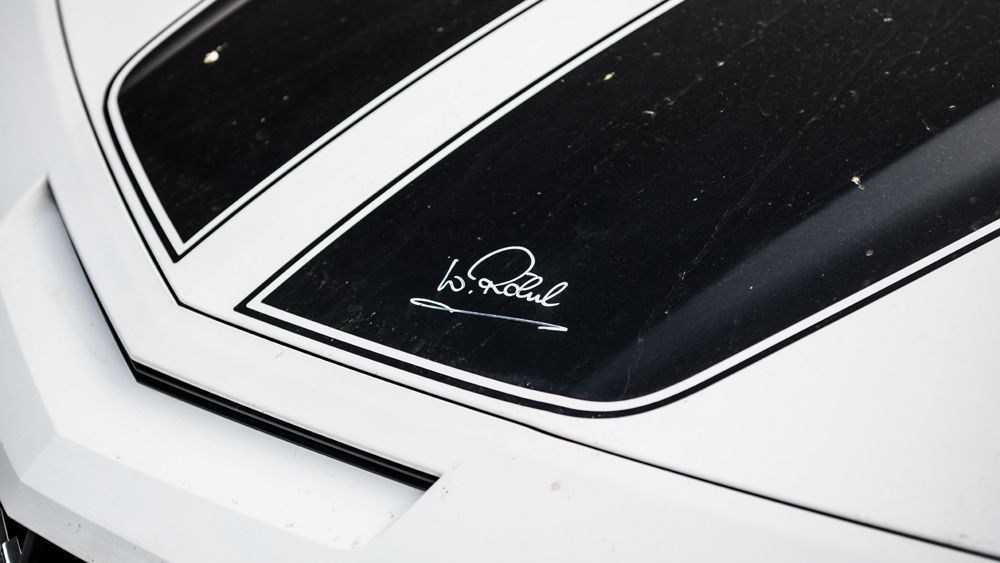 Weiße Motorhaube mit schwarzen Rennstreifen und einer Unterschrift.