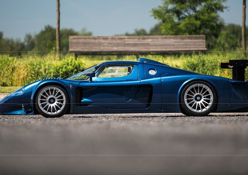 Blauer Maserati MC12 auf Asphalt geparkt, Seitenansicht. Silberne Räder, schwarzer Spoiler, sonnige Außeneinstellung.