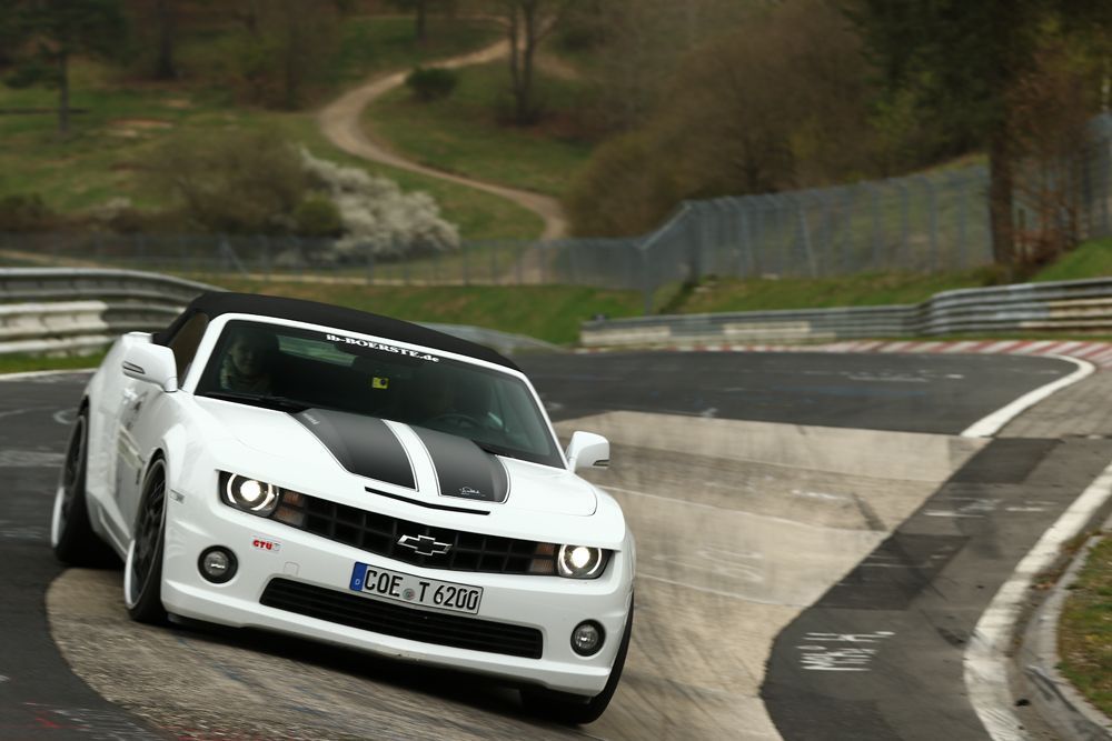 Weißes Chevrolet Camaro-Cabrio rast mit schwarzen Rennstreifen auf der Motorhaube um eine Rennstreckenkurve.