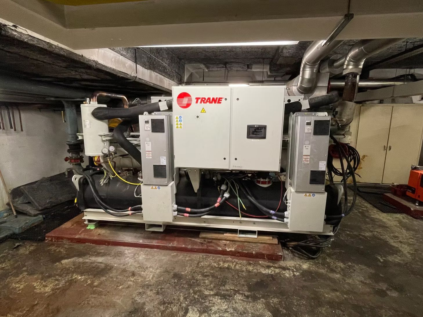 Gros appareil CVC Trane dans un local technique au sous-sol, avec des tuyaux, des conduits et un sol en béton.