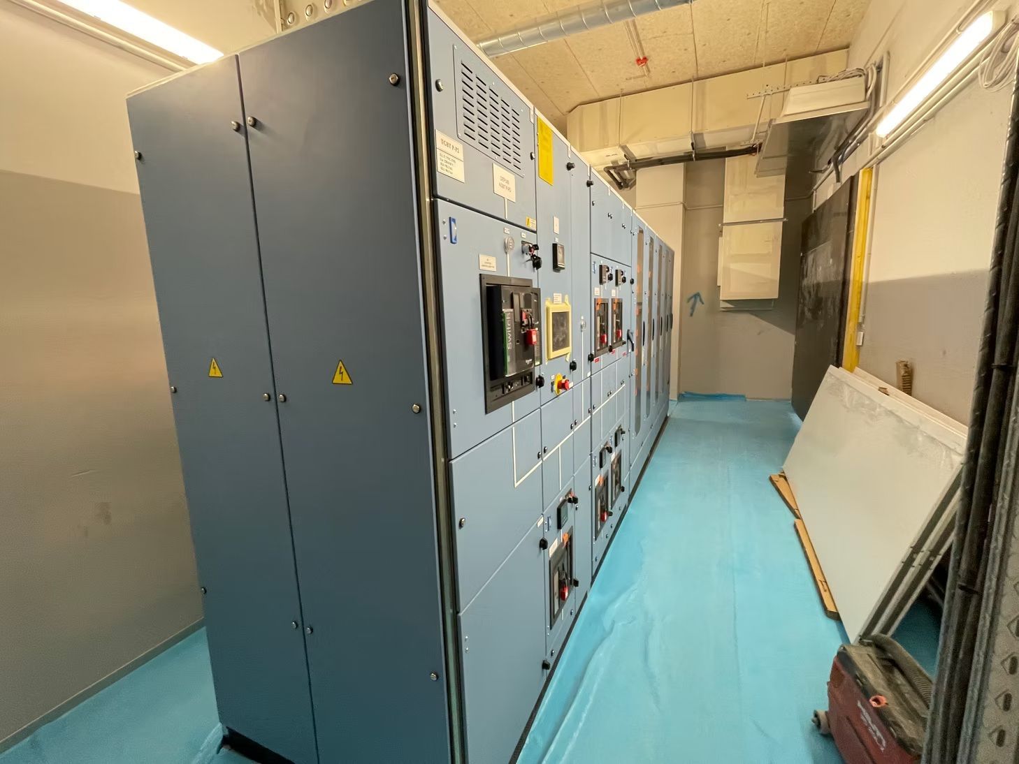 Salle électrique industrielle avec des panneaux de commande bleu-gris alignés le long d’un couloir étroit.