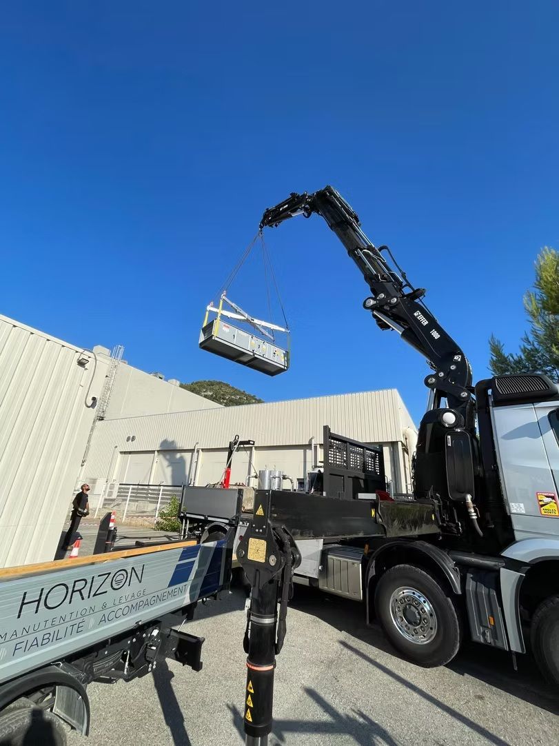 Un camion-grue soulève une charge suspendue à côté d’un bâtiment, sous un ciel bleu dégagé