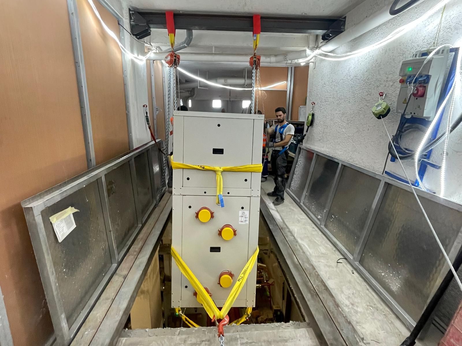 Machine industrielle blanche sanglée dans un couloir étroit réservé aux équipements, avec des ouvriers visibles à l’arrière-plan
