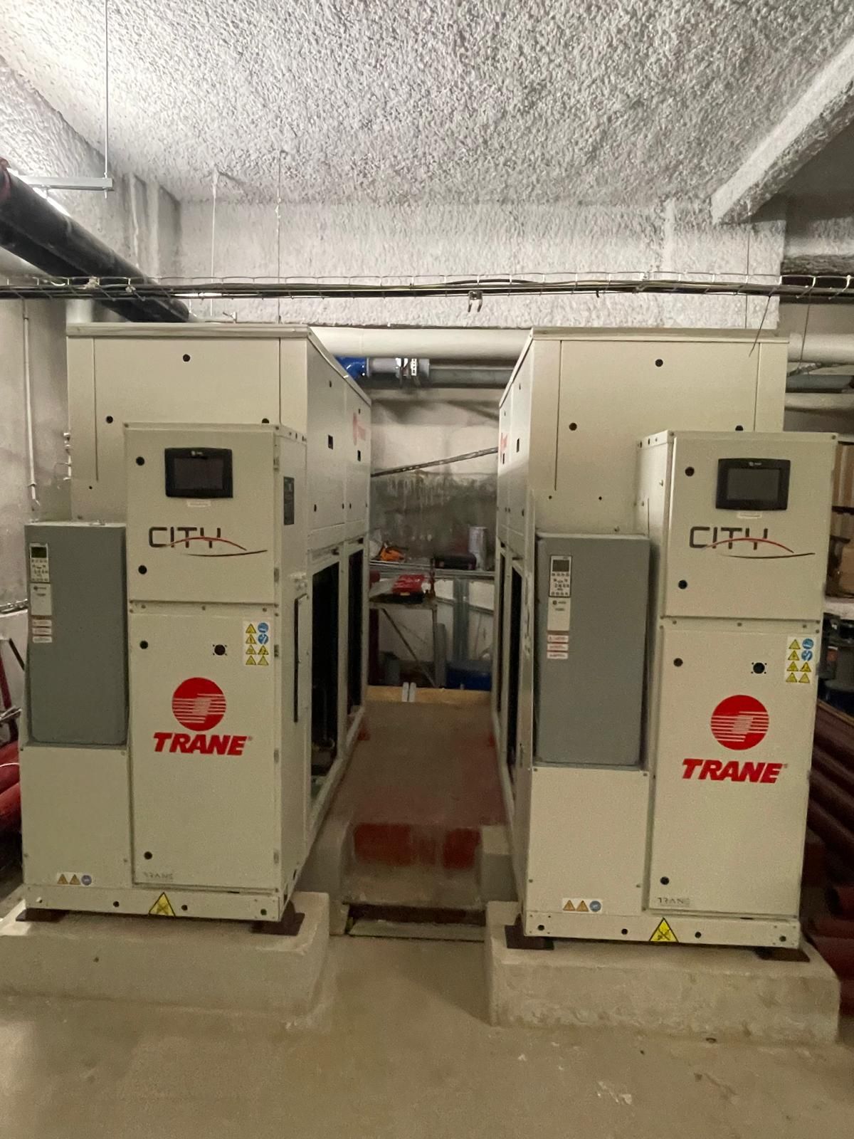 Deux grandes unités CVC Trane blanches dans un local technique en béton, avec une allée étroite entre elles