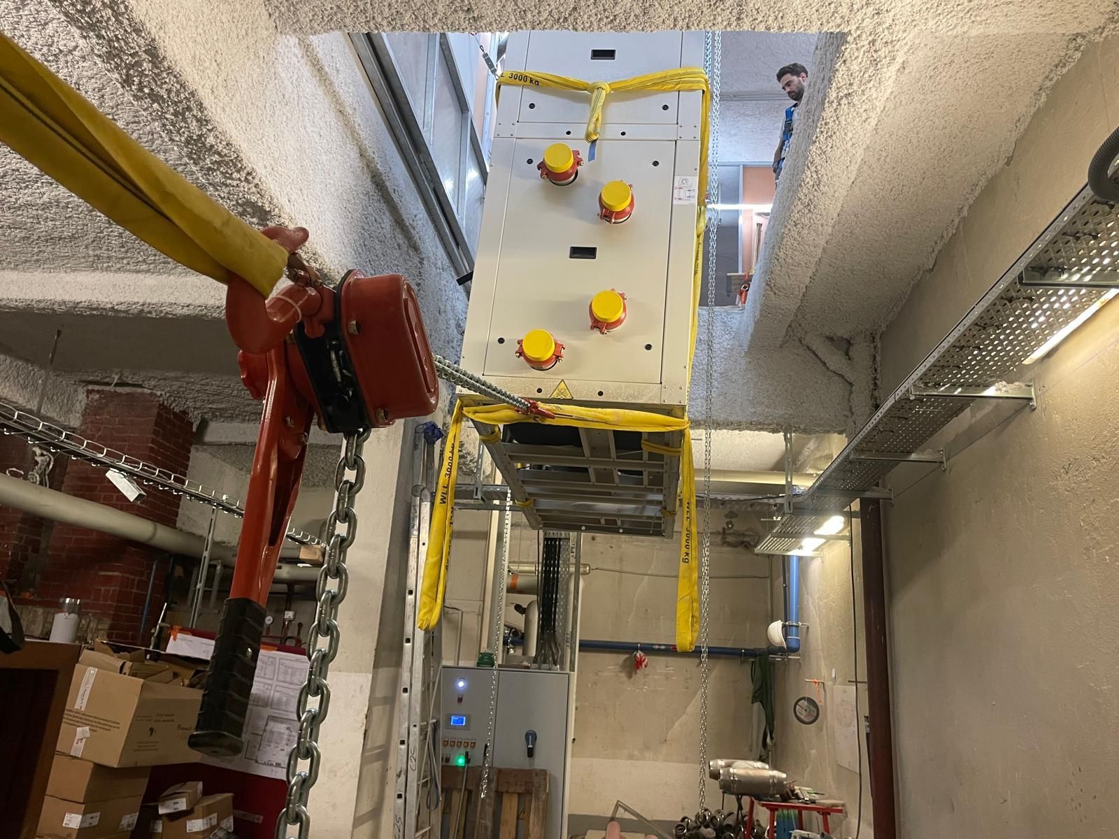 Plateforme de travail industrielle suspendue à une grue dans une gaine technique étroite, avec des garde-corps jaunes et un bras de levage rouge