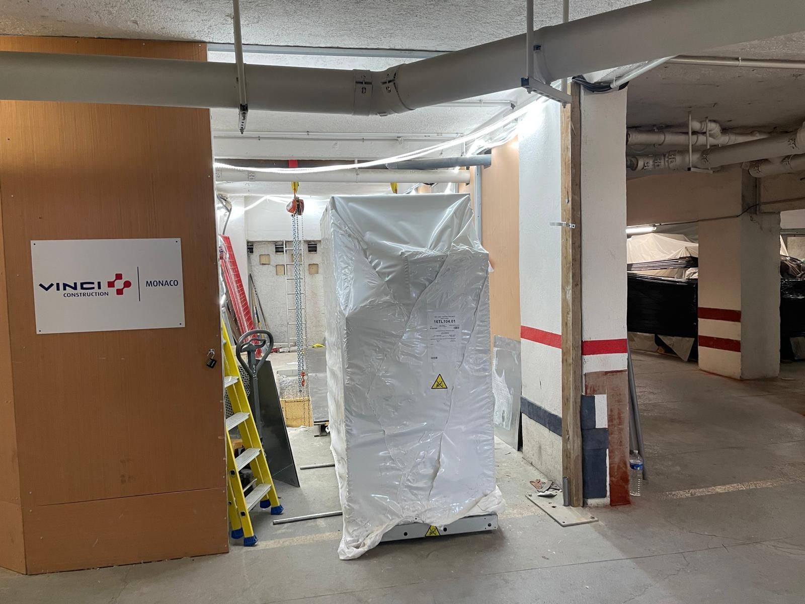 Zone de travaux avec du matériel emballé, une échelle et des colonnes en béton peint dans un parking souterrain inachevé