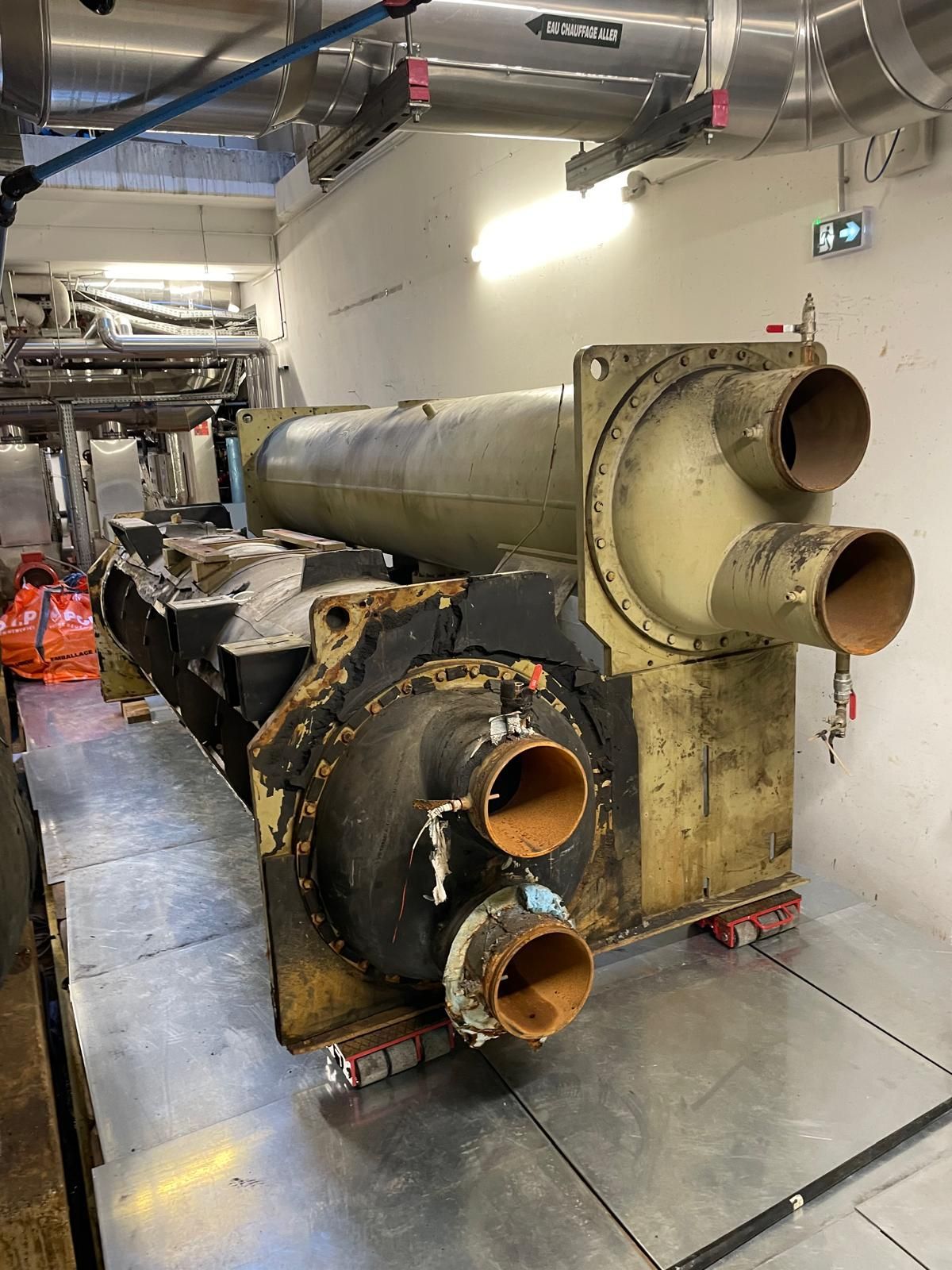 De grosses machines industrielles dans une galerie de maintenance, avec des tuyaux rouillés et des ouvriers portant des gilets orange à l’arrière-plan