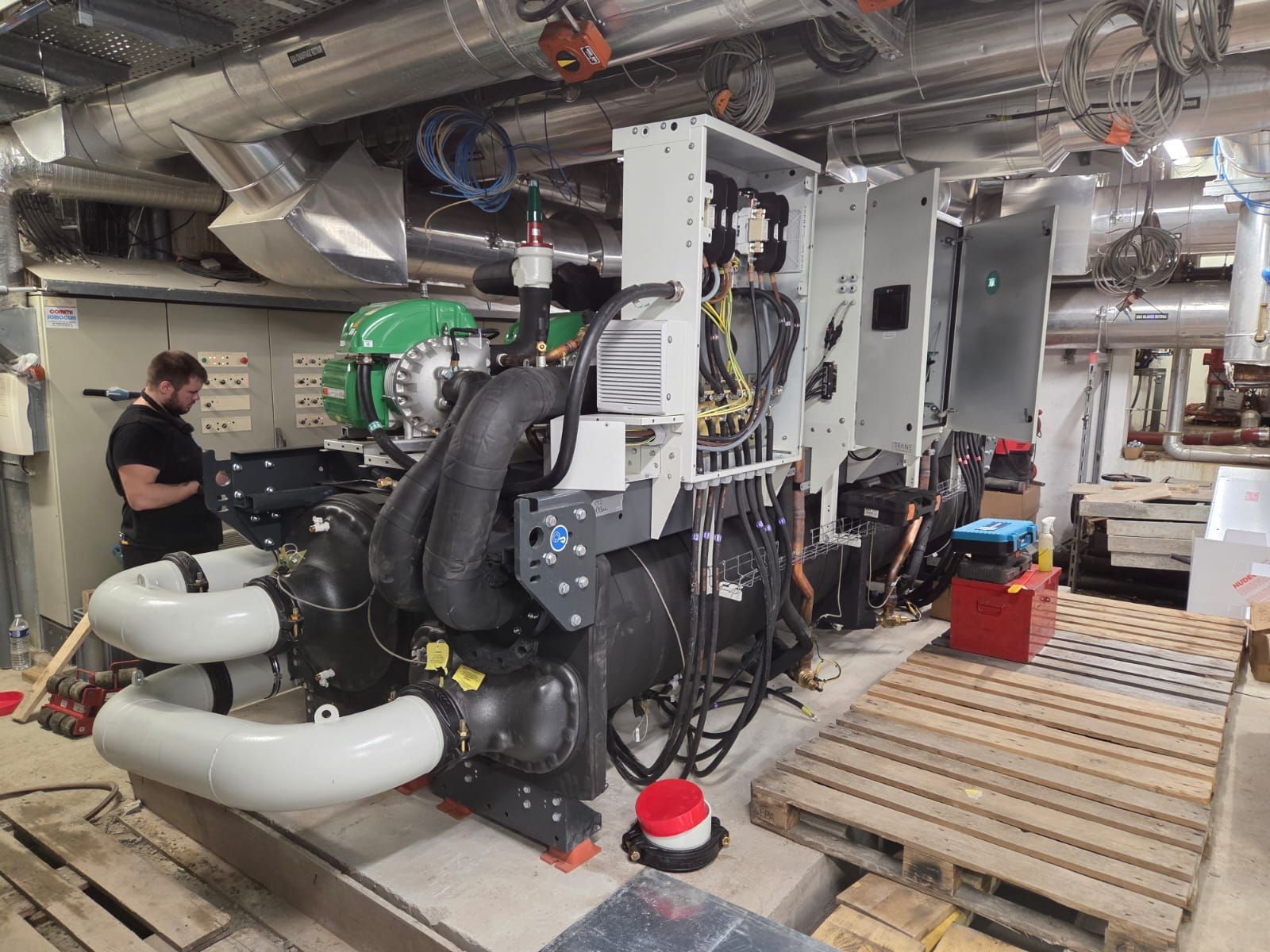 Salle des machines industrielles avec de grosses machines, des tuyaux et des conduits ; un ouvrier se tient à gauche, près des équipements.