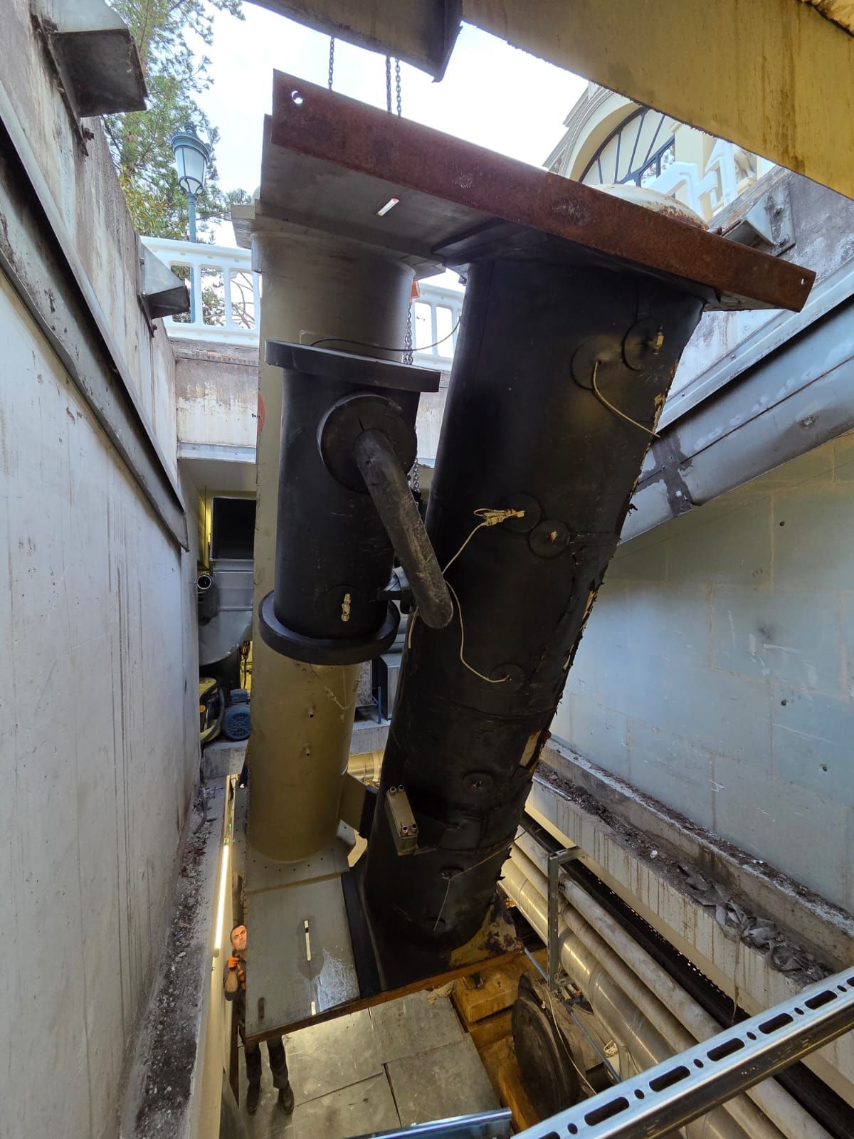 Grosses canalisations et conduits noirs dans une gaine technique étroite aux murs en béton, avec une verrière au-dessus