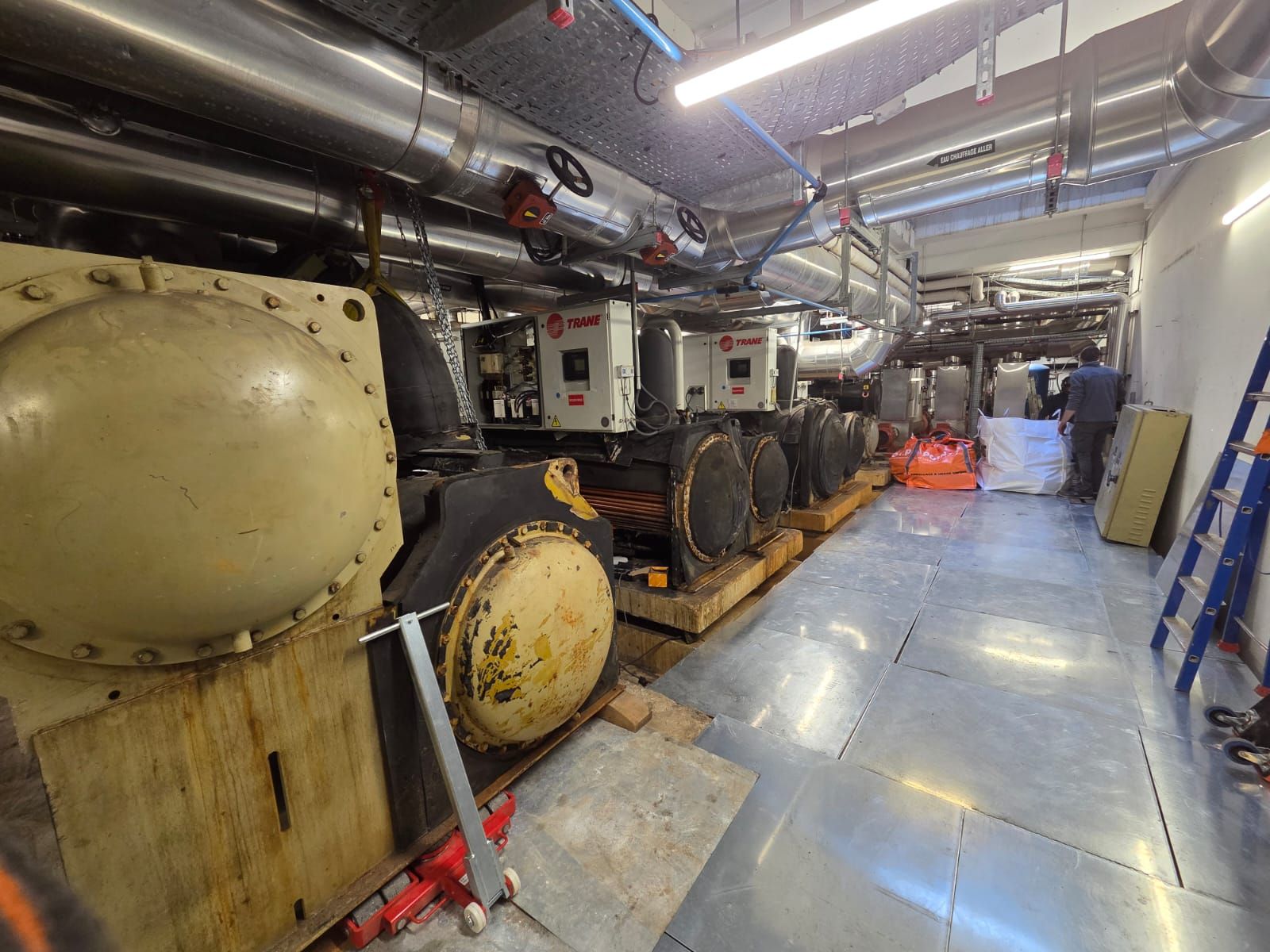 Des machines industrielles et des tuyauteries dans un couloir technique étroit, avec un sol bleu et des éclairages au plafond.