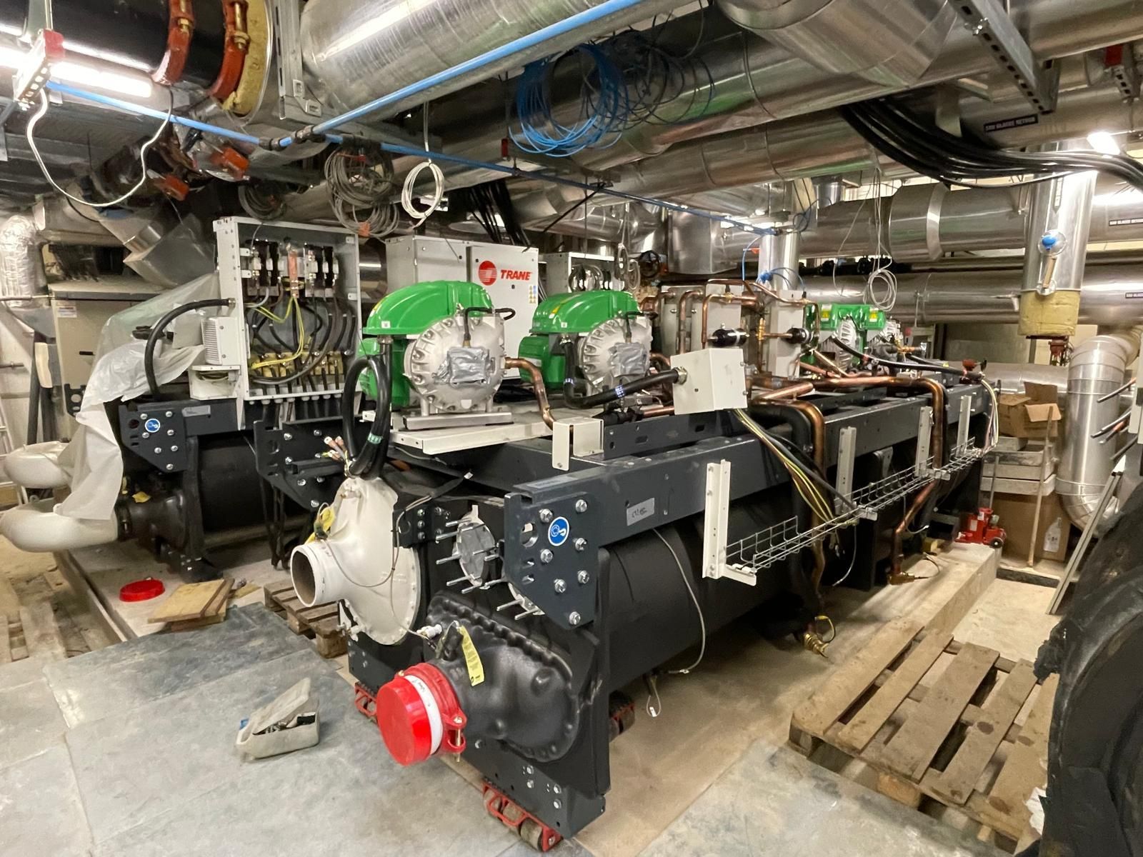 Grande salle des machines industrielle avec deux générateurs verts et noirs, des tuyaux au plafond et des palettes en bois au sol