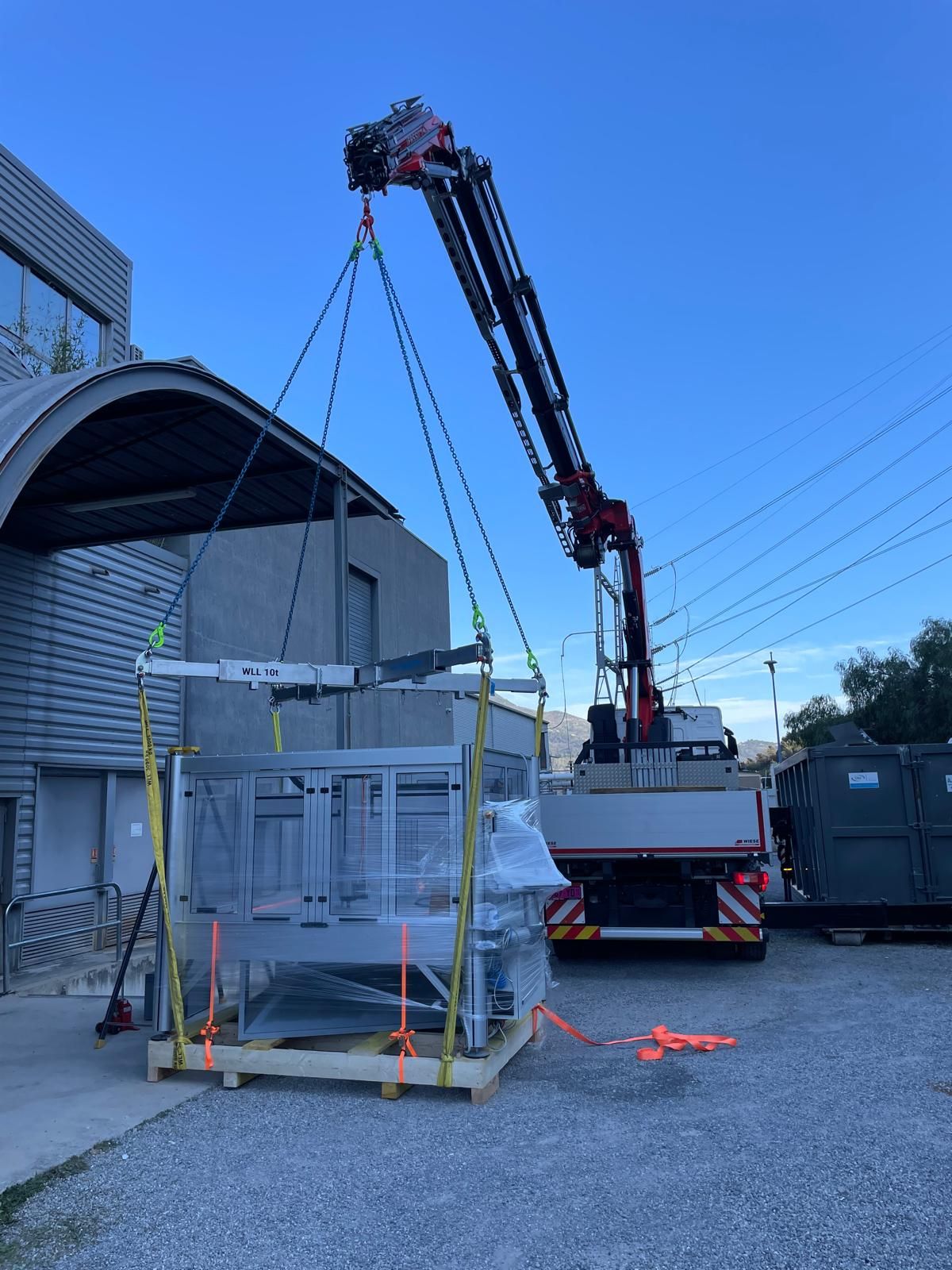 Grue soulevant une palette emballée à côté d’un bâtiment sous un ciel bleu dégagé