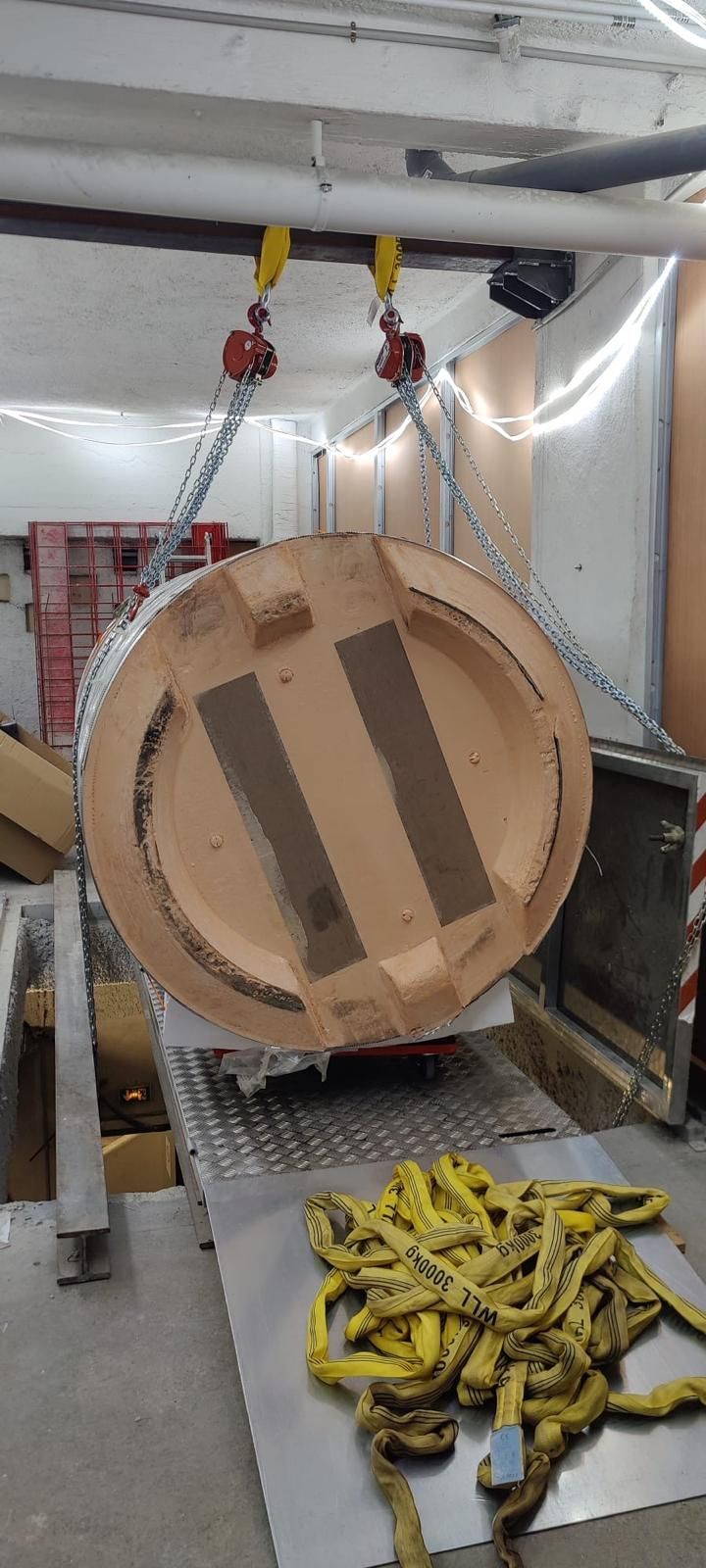 Grande bobine en bois suspendue dans un camion de marchandises, avec des sangles jaunes et des chaînes au sol.