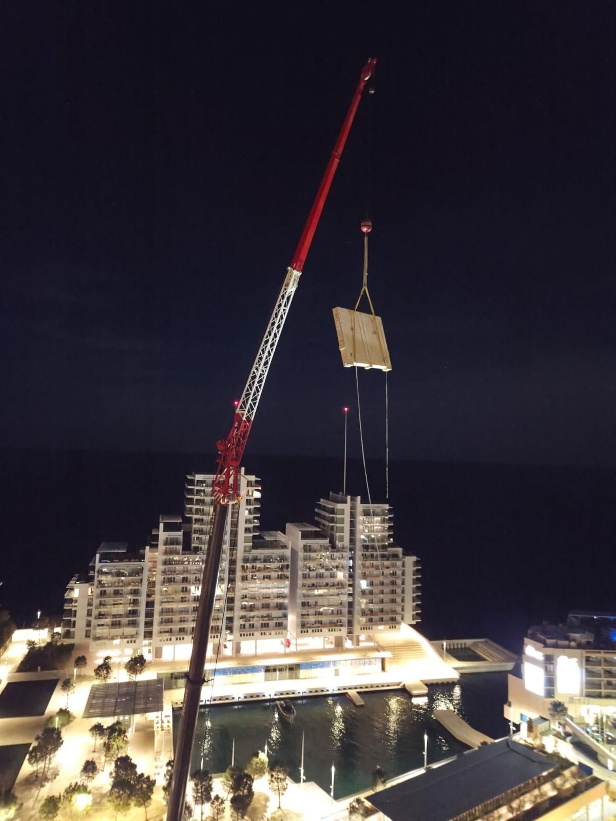 Une grue de nuit soulève un grand panneau rectangulaire au-dessus d’un bâtiment éclairé au bord de l’eau, à la tombée du jour