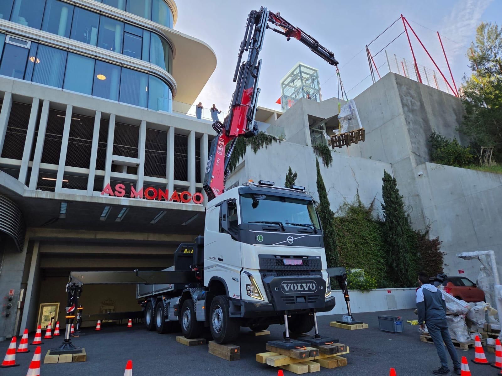 Camion-grue Volvo blanc soulevant des matériaux près d’un bâtiment moderne en construction, avec des ouvriers à proximité