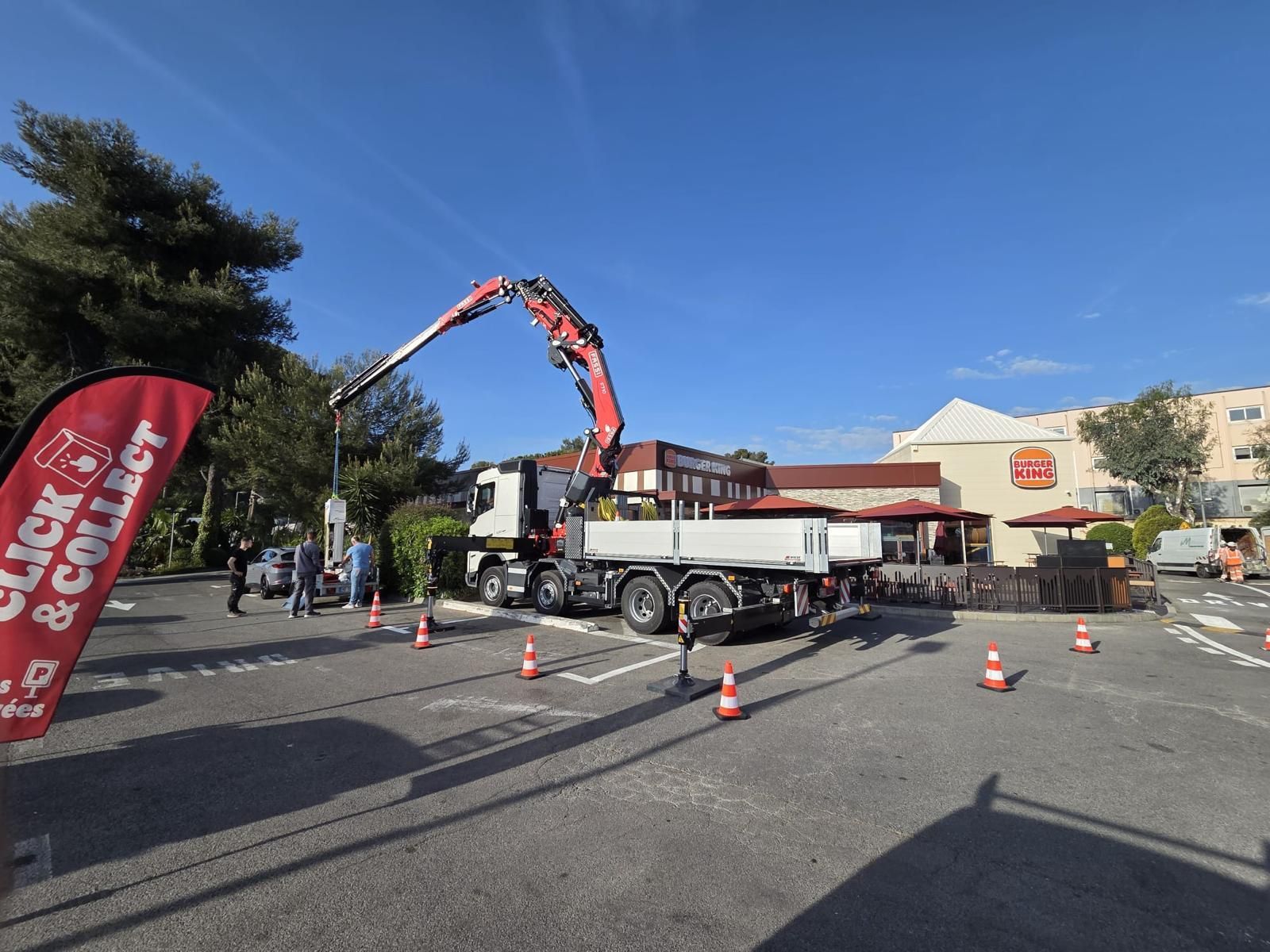 Grue montée sur camion soulevant des matériaux sur un parking, avec des cônes et une banderole rouge à proximité.