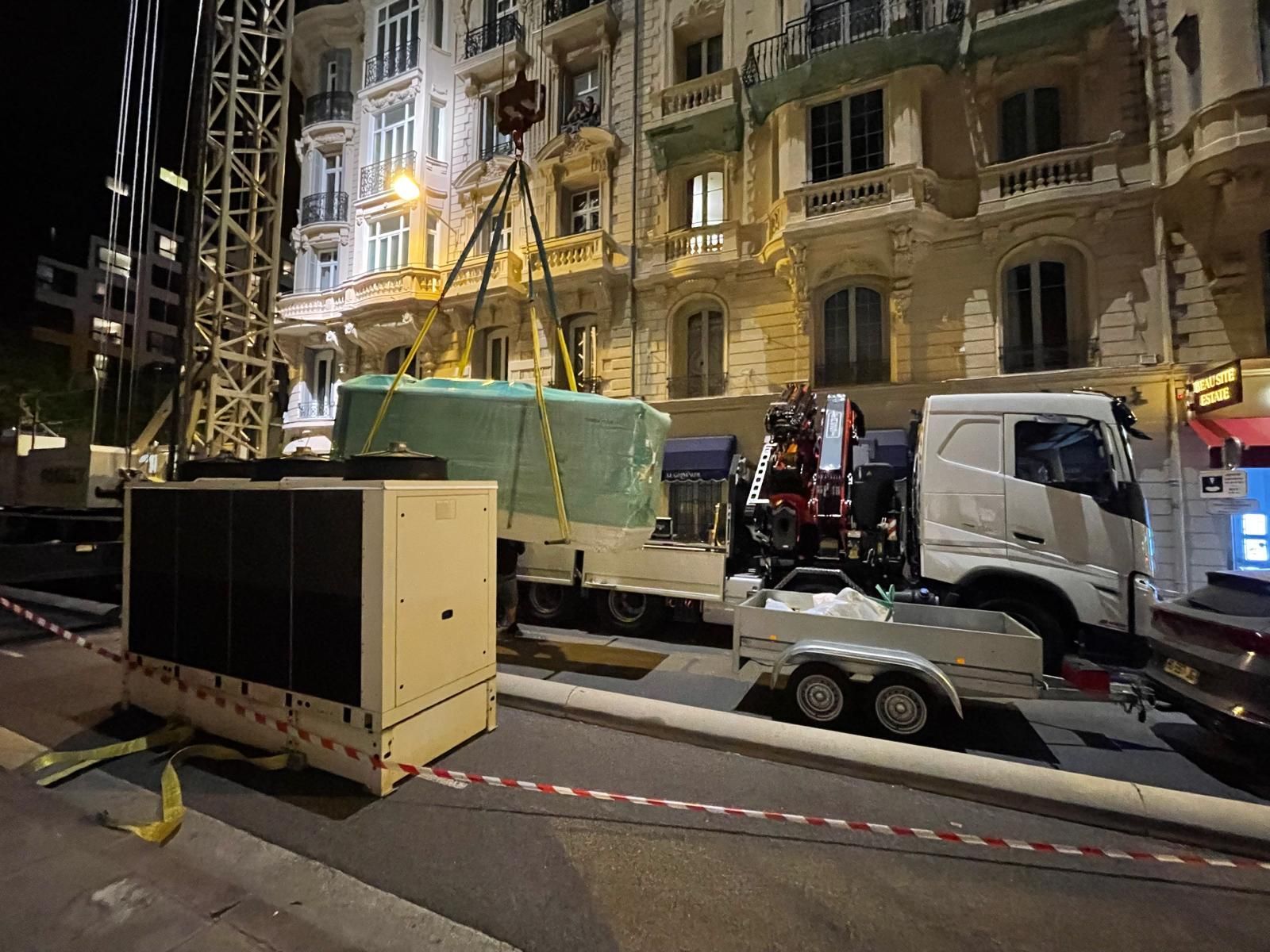 Scène de rue nocturne avec une grue, des camions et du matériel de chantier devant un bâtiment orné