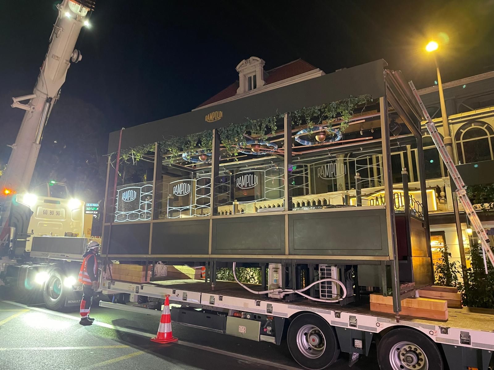 Structure en forme de maison transportée de nuit sur un camion, entourée de cônes et accompagnée d’une grue.
