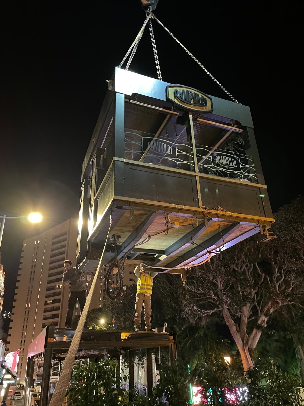 Une grue soulève la charpente d’un petit bâtiment de nuit, avec des lumières et des arbres en contrebas