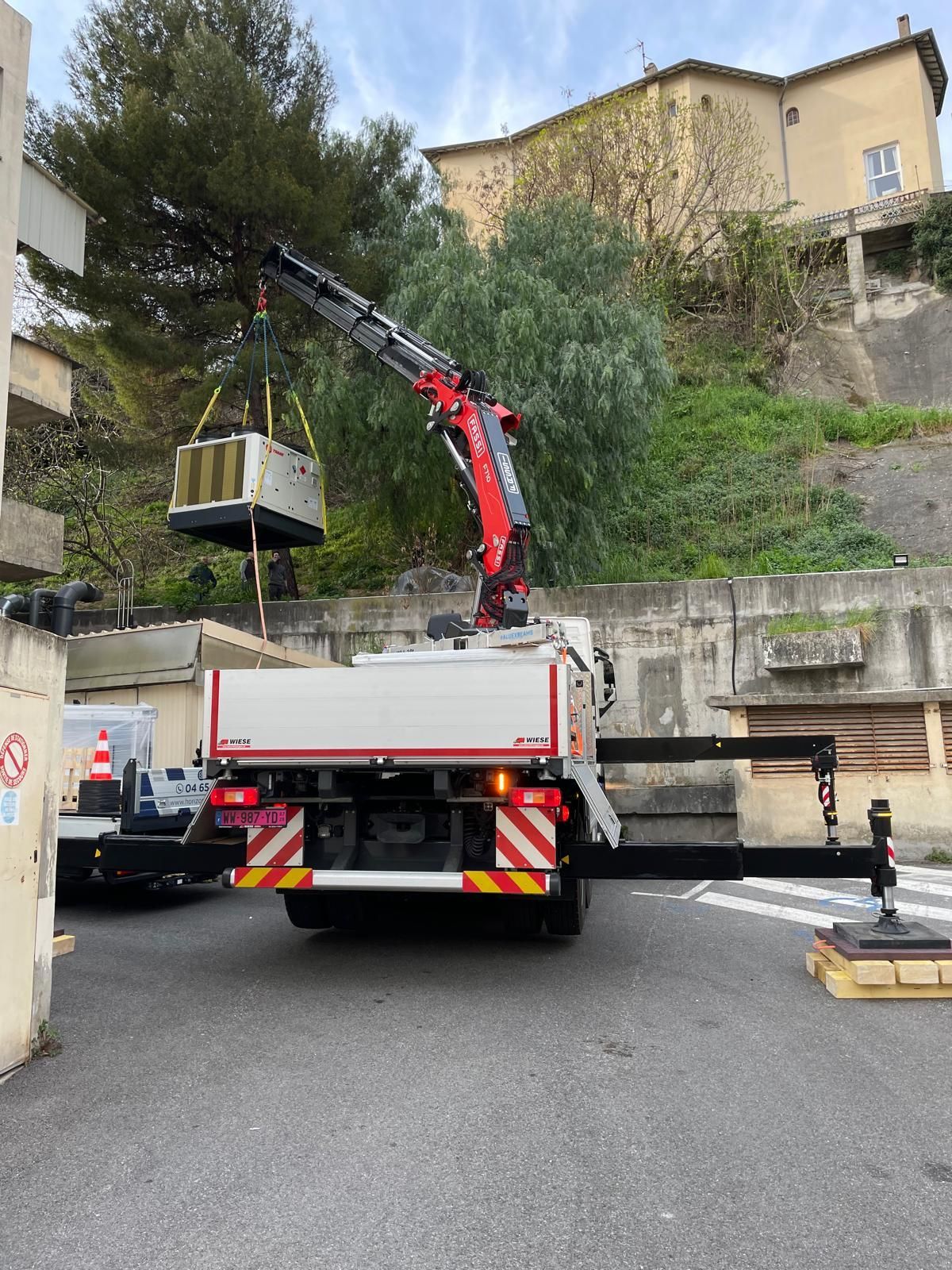 Un camion-grue soulève une benne métallique sur une rue résidentielle en pente bordée de murs en pierre et de maisons