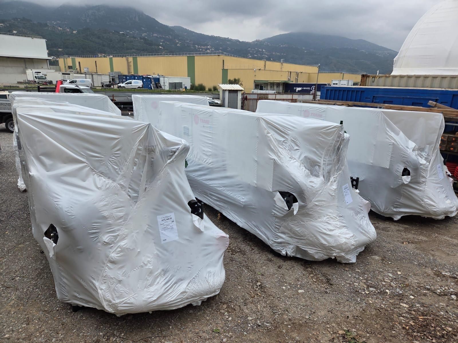 Des machines emballées sous film plastique sur des palettes dans une cour industrielle en plein air, avec des montagnes en arrière-plan