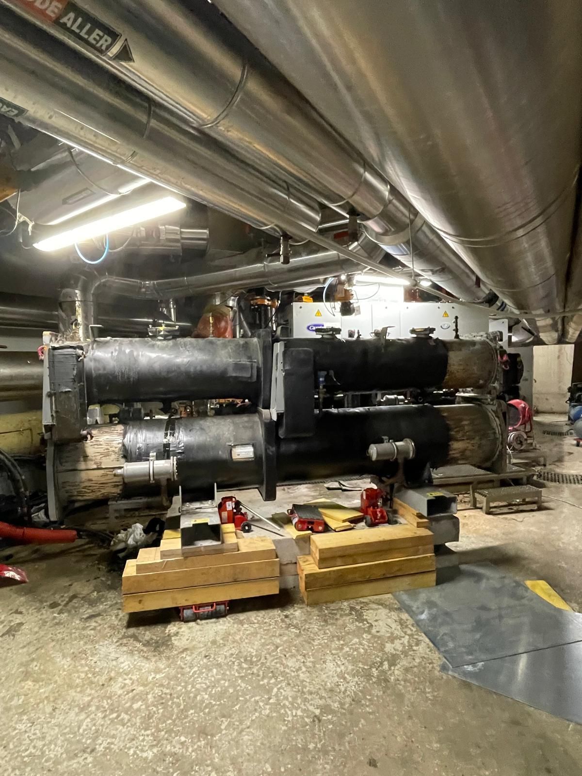 Des machines industrielles dans une pièce en sous-sol, avec des tuyaux au plafond et des palettes de fournitures au sol