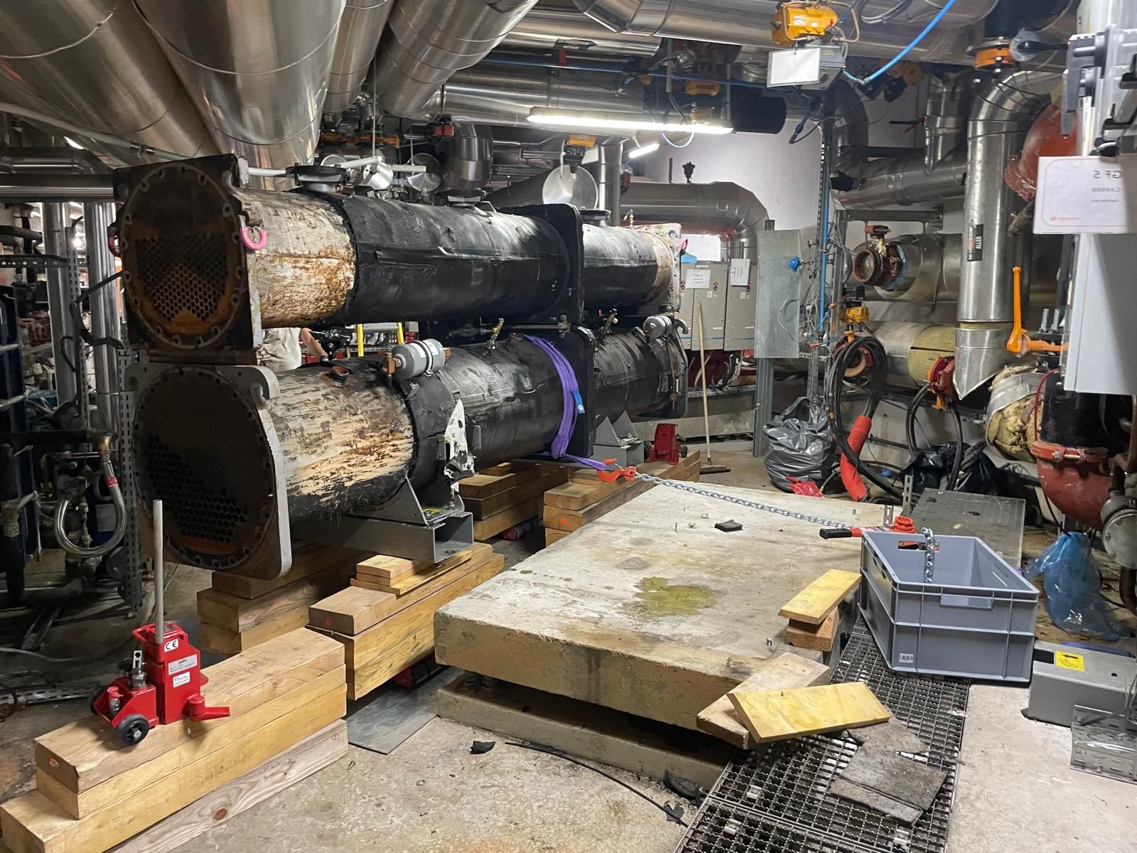 Local industriel avec des tuyaux, des conduits et de l’isolation, ainsi que des palettes en bois et des outils au sol.
