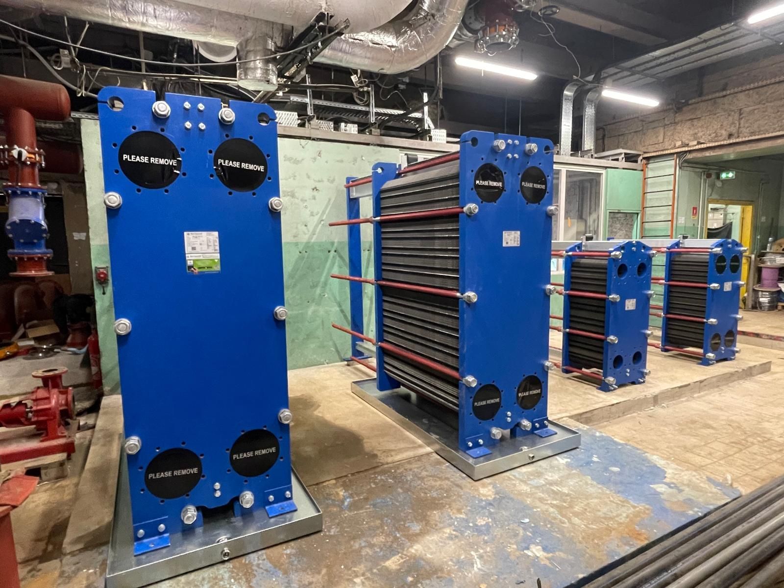 Des machines industrielles bleues équipées de rouleaux rouges, disposées dans un atelier d’usine sous des tuyaux apparents.
