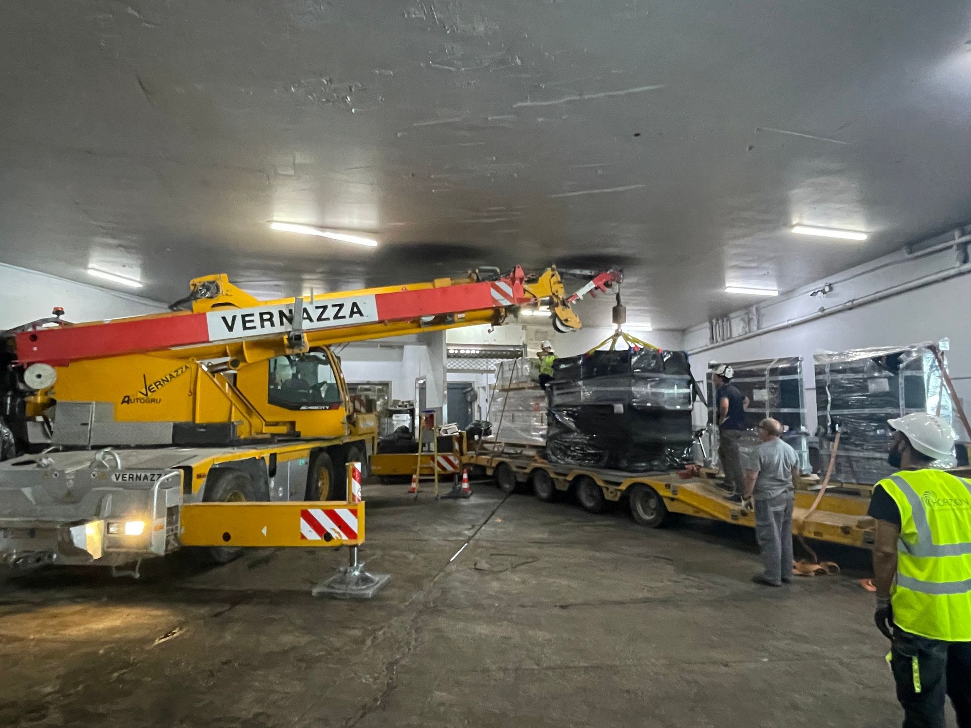 Une grue charge un grand véhicule noir sur une remorque dans un garage couvert