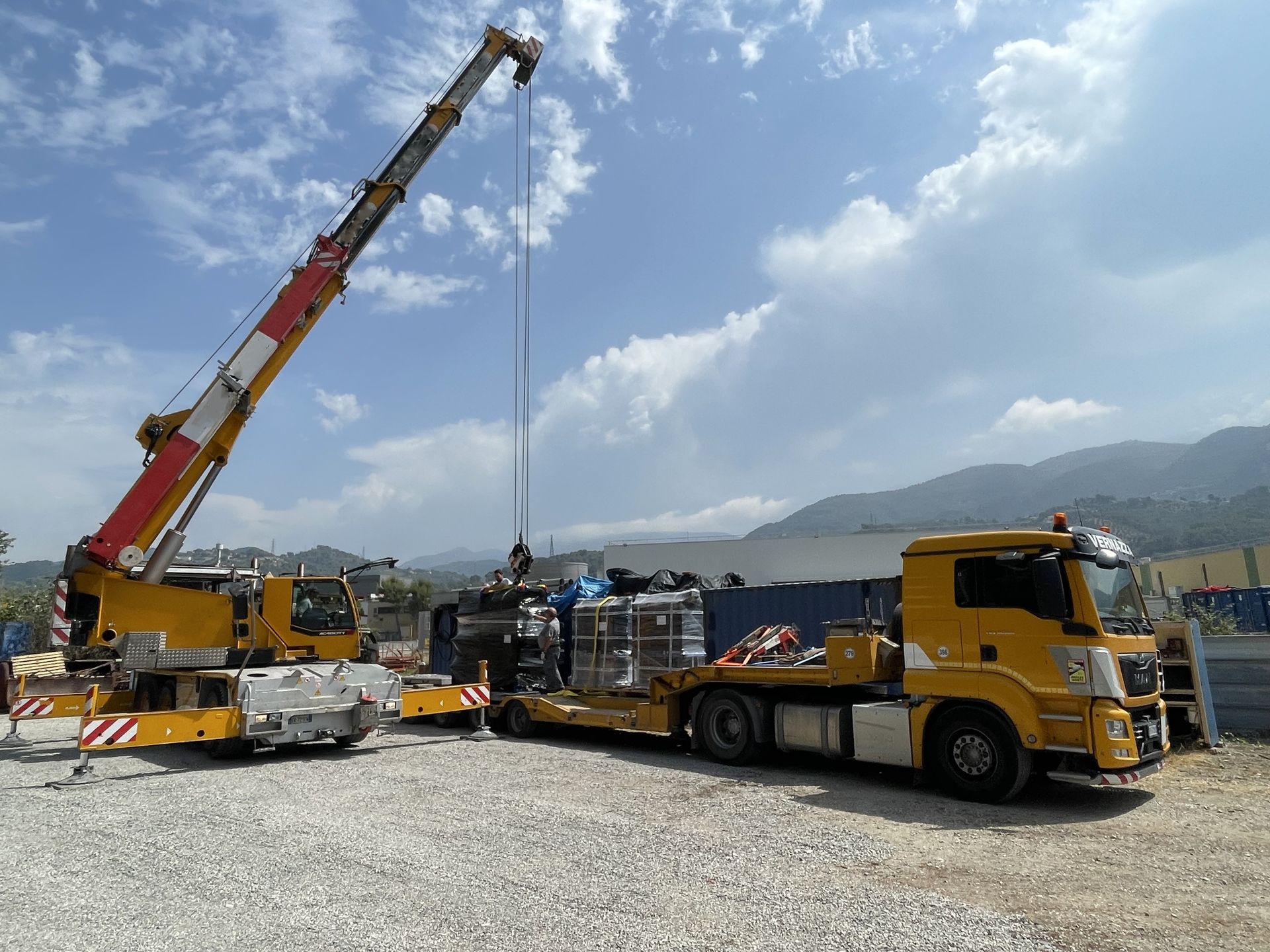 Une grue charge du matériel sur un camion à plateau jaune dans un terrain en gravier, sous un ciel nuageux