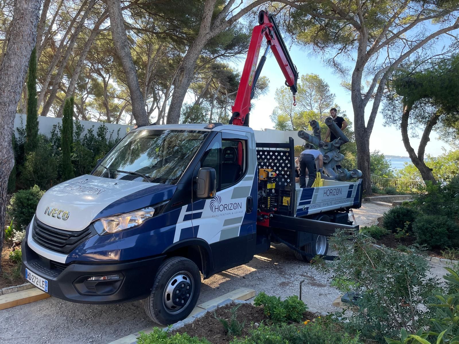 Camion utilitaire bleu avec une grue déployée, garé sur un chemin de gravier au milieu des arbres et des buissons