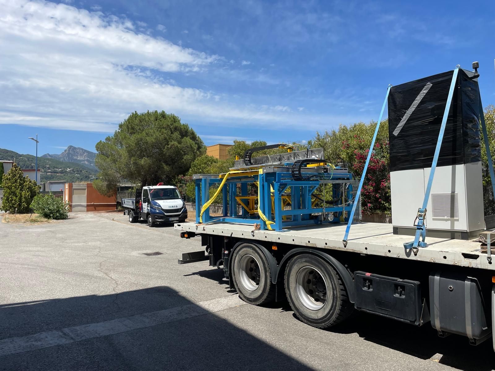 Un camion à plateau transporte du matériel métallique bleu sur un parking ensoleillé, près de bâtiments et de montagnes