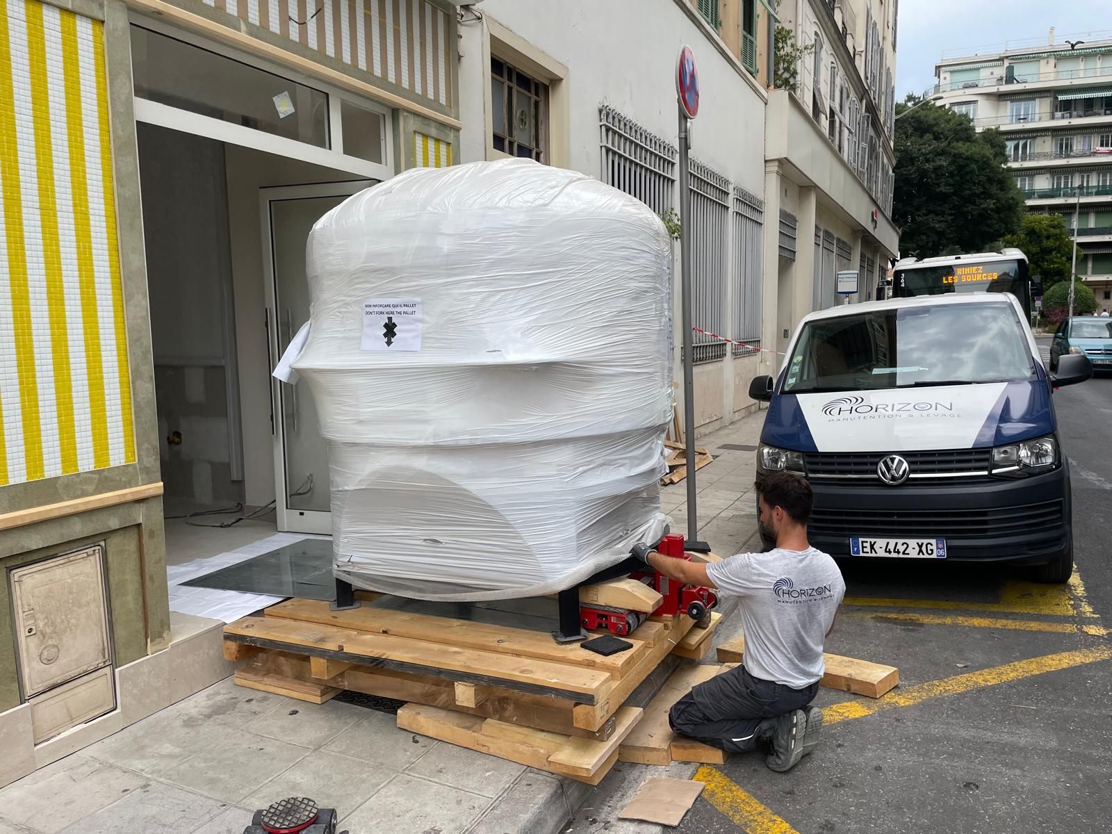 Une personne est agenouillée à côté d’une palette emballée, sur un trottoir bordé de voitures garées et d’un bâtiment.